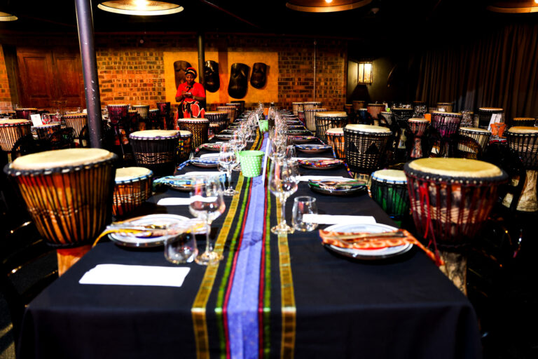 Long candlelit dinner table set for a private celebration, lined with African djembe drums